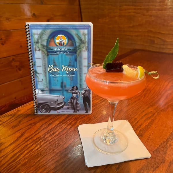 A vibrant pink cocktail with mint and lemon garnish sits next to a Copacabana Cuban Cafe bar menu.