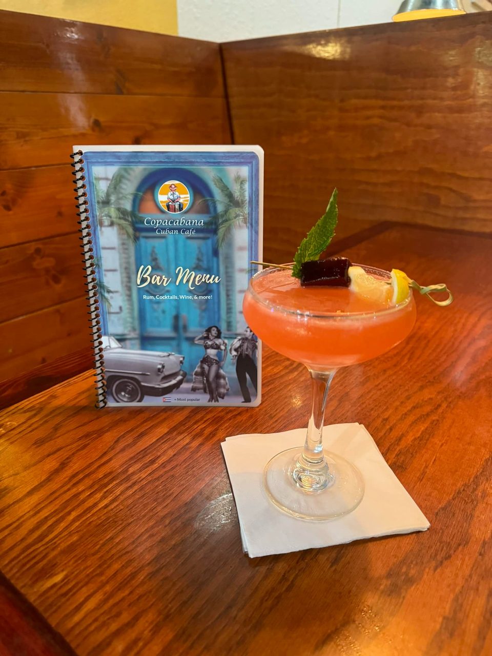 A vibrant pink cocktail with mint and lemon garnish sits next to a Copacabana Cuban Cafe bar menu.