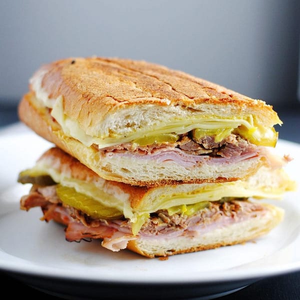 A Cuban sandwich, cut in half and stacked, revealing ham, roasted pork, Swiss cheese, and pickles.